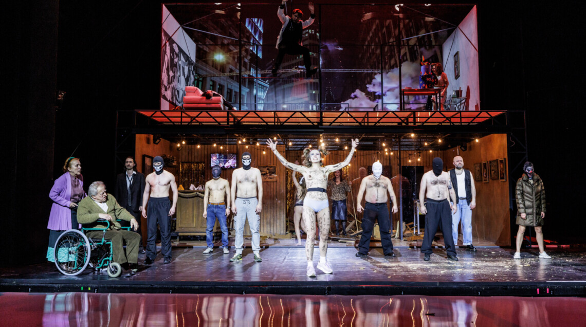 Grupa osób ustawionych na scenie, w centrum kobieta w samych majtkach, na całym ciele ma wypisane na czarno napisy, za nią stoją różne osoby, głównie mężczyźni bez koszulek w maskach, z lewej strony mężczyzna siedzący na wózku inwalidzkim oraz kobieta pchająca wózek