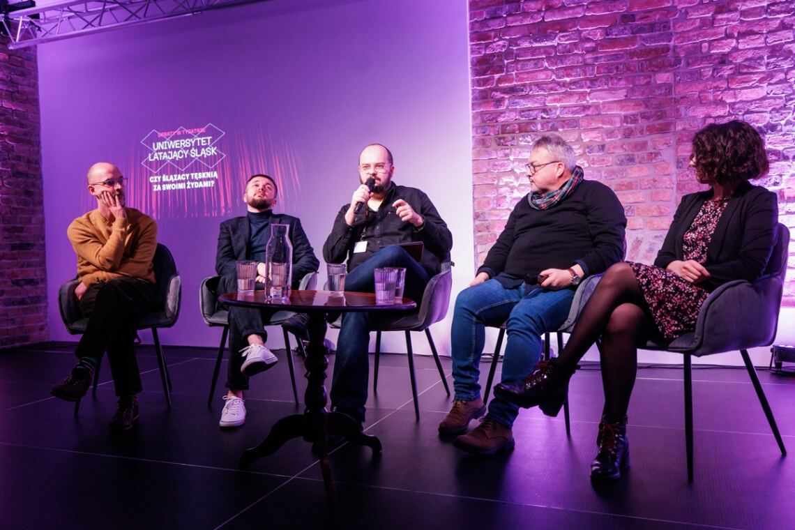 Na scenie siedzi czterech mężczyzn oraz jedna kobieta, siedzący w środku mężczyzna prowadzący spotkanie przemawia do mikrofonu, przybliżenie