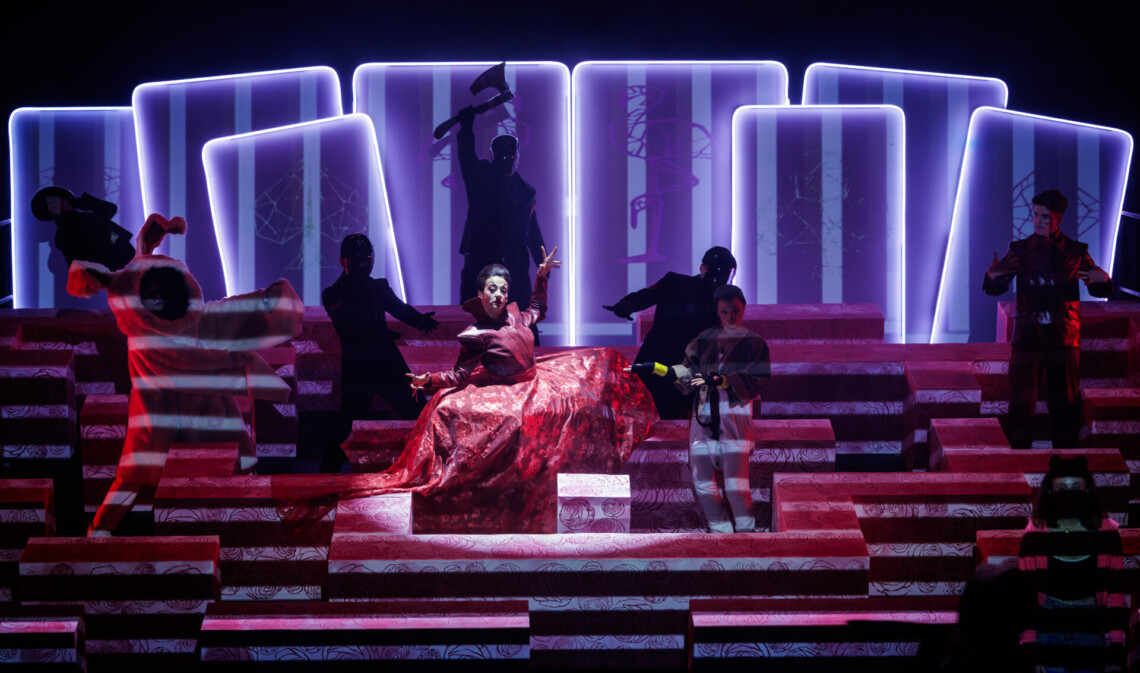 Niski, czerwony labirynt, w którym znajdują się różne postacie, w centrum kobieta w czerwonej sukni, obok biały królik, w tle mężczyzna z toporem, na końcu labiryntu stoją karty do gry