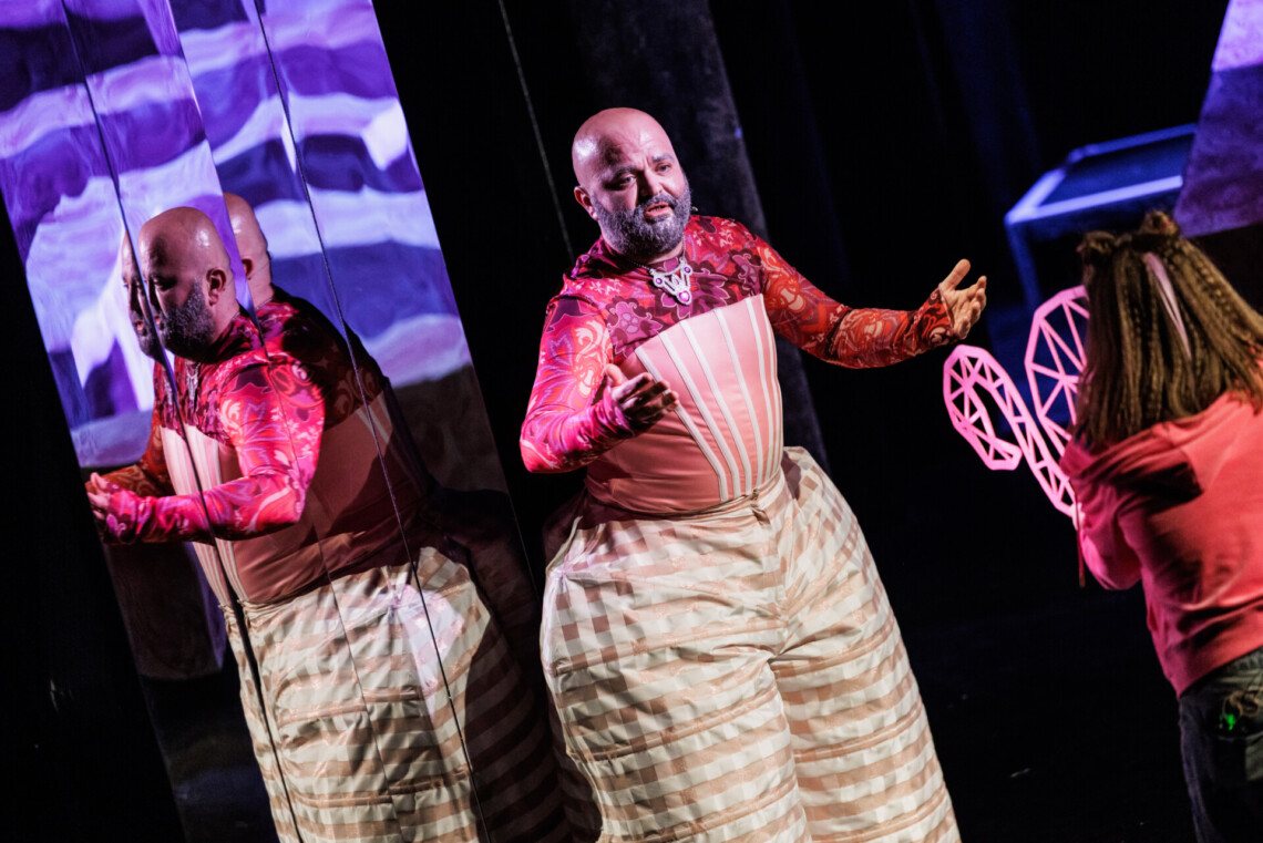 Masywna postać w jasnoróżowym kostiumie z szerokimi, wypchanymi spodniami w kratkę, mężczyzna stoi z rozłożonymi rękami, odbija się w lustrzanej ścianie za nim, przed nim stoi dziewczyna, która widoczna jest od tyłu