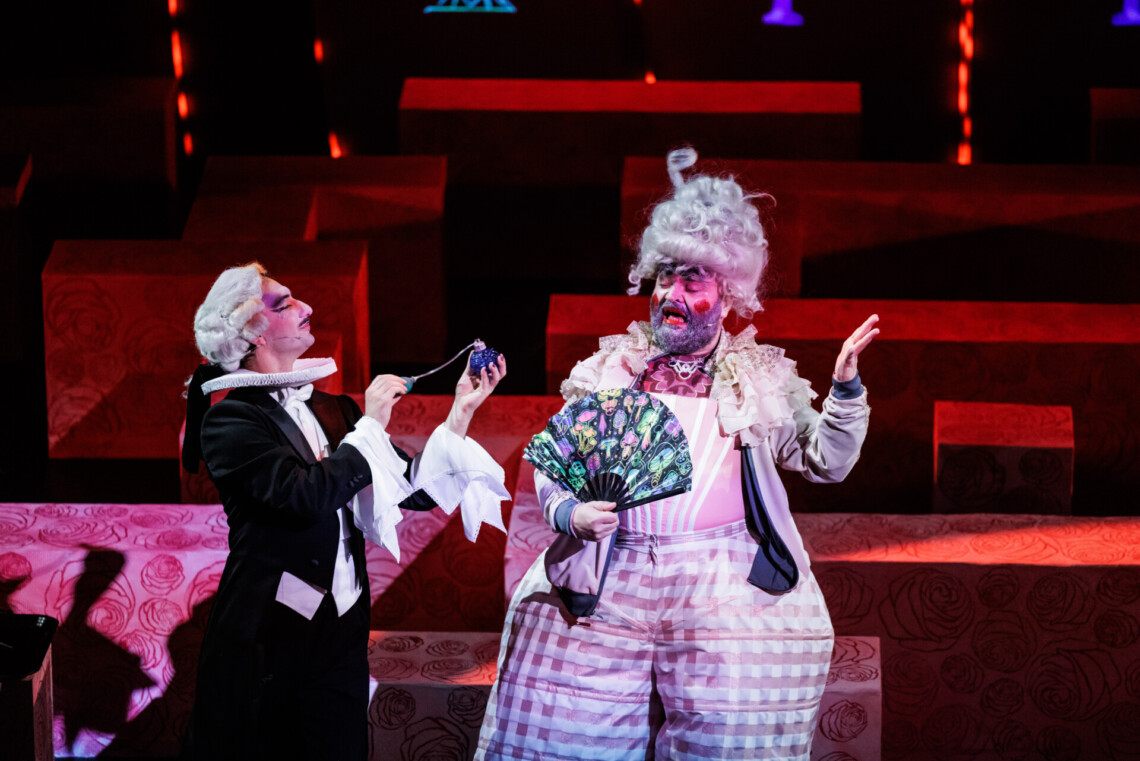 Mężczyzna w czarnym fraku psika perfumami z pompki w stronę drugiej osoby z peruką, która z zamkniętymi oczami lekko się odsuwa, stojąc na tle czerwonego labiryntu