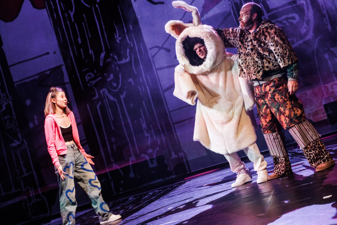 Z lewej strony stoi ubrana współcześnie dziewczyna, z prawej stoi królik oraz mężczyzna w kurteczce w cętki jako Kot, Królik ma duży, biały, pluszowy kostium z długimi uszami, przez którego otwór na twarz widać głowę aktora