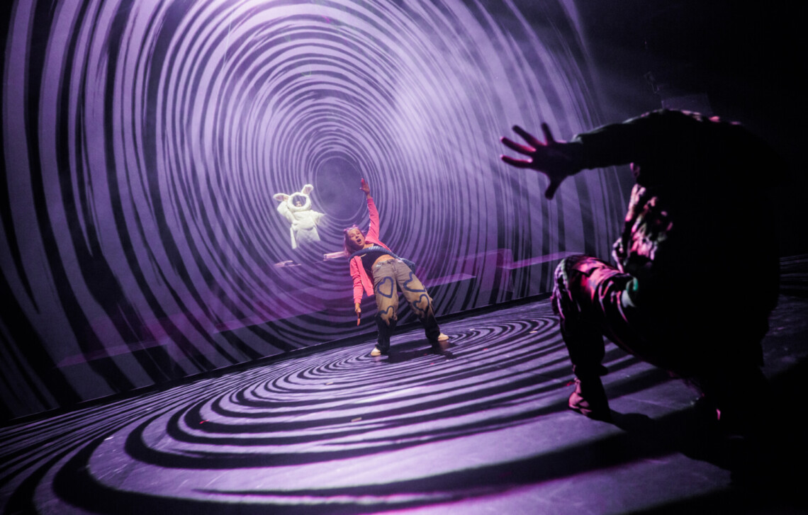 W centrum stoi dziewczyna pochylona do tyłu z jedną ręką uniesioną w górę, za nią znajduje się spiralna, fioletowa ściana, wygląda jakby dziewczyna była wciągana przez wir, za spiralną ścianką widoczna jest postać białego królika, a przed dziewczyną z boku w półmroku widoczna jest sylwetka postaci wyciągającej ku niej dłoń