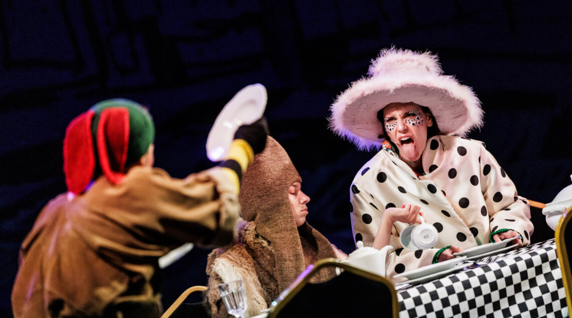 Postaci za stołem z obrusem w biało-czarną kratkę, wyraźna postać osoby z dużym kapeluszem w białek kurtce w czarne grochy, ma wyciągnięty do przodu język