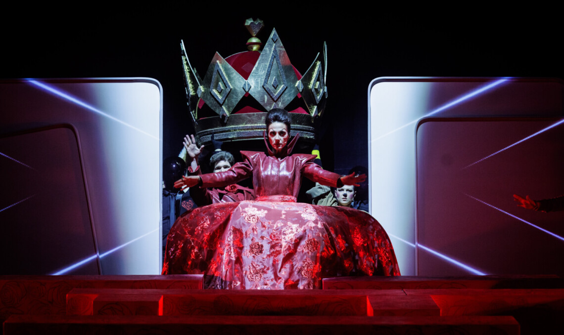 W centrum kobieta w czerwonej, rozłożystej sukni, z wyciągniętymi w bok rękami, za nią wznosi się wielka, złoto-czarna korona podtrzymywana przez dwie osoby, po bokach jasne panele, tło mroczne, zdjęcie jest symetryczne