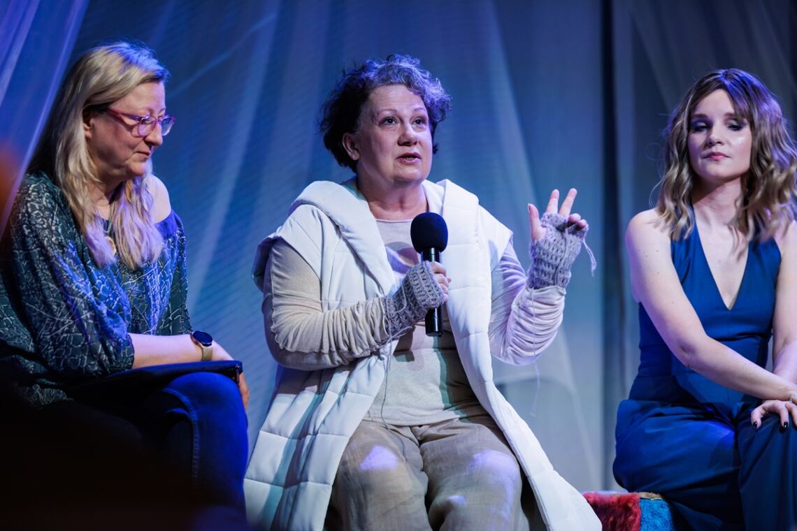Na scenie siedzą trzy kobiety: Agata Kurzak, Violetta Smolińska oraz Agnieszka Radzikowska. Violetta trzyma w prawej dłoni mikrofon do którego przemawia, lewa dłoń z wyciągniętymi trzema palcami uniesiona do góry.