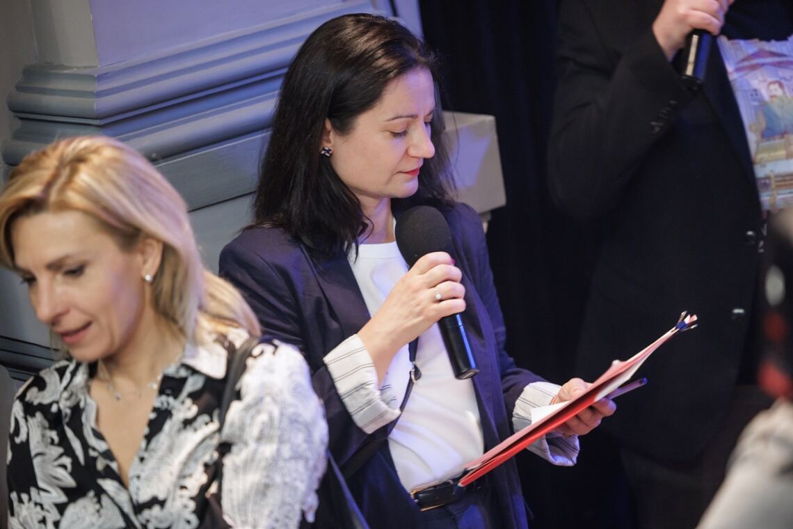 Prowadząca konferencję kobieta stoi z mikrofonem w prawej dłoni. W lewej dłoni trzyma teczkę, na którą spogląda.
