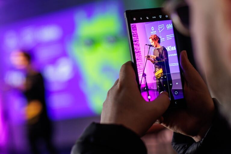 Postać robiąca zdjęcie kobiecie przemawiającej na scenie. Widok na dłonie i smartphone.