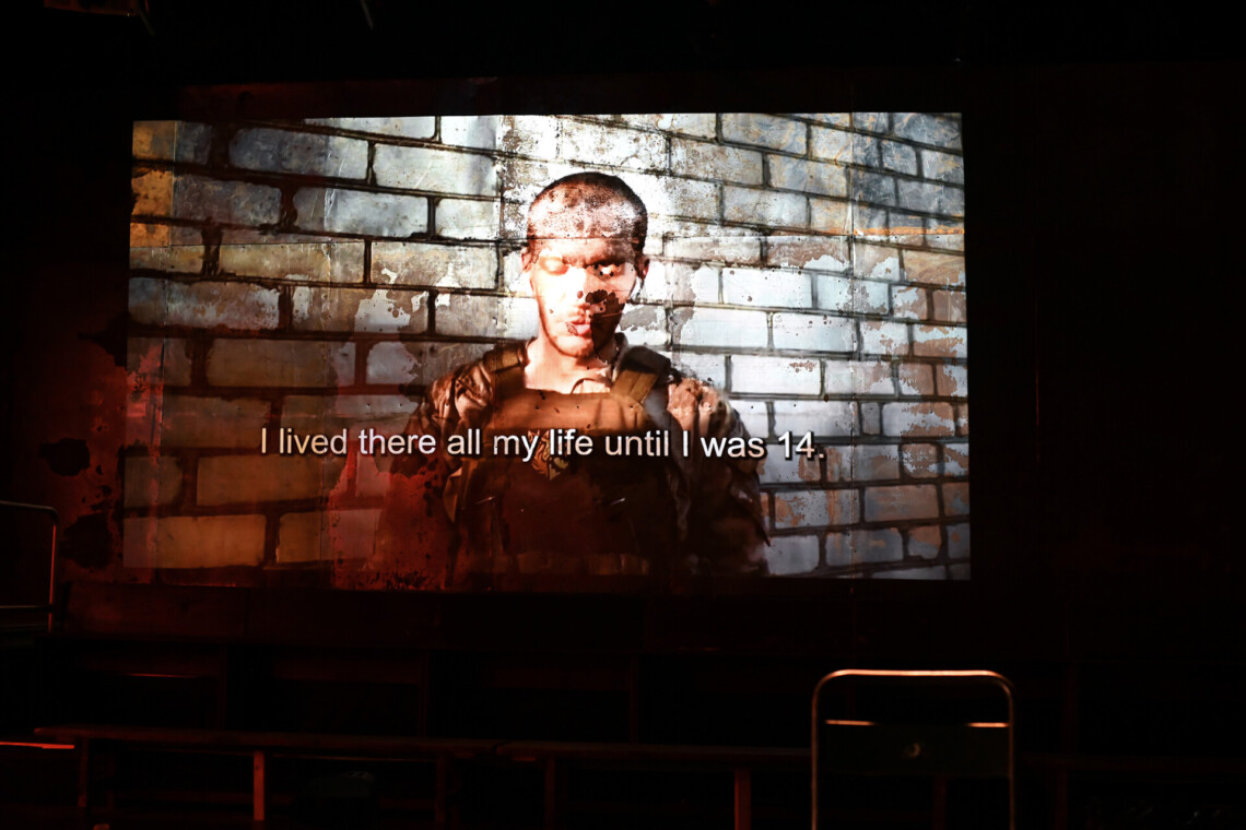 A film with subtitles is being screened on the wall, with a man in a military uniform, his eyes are closed