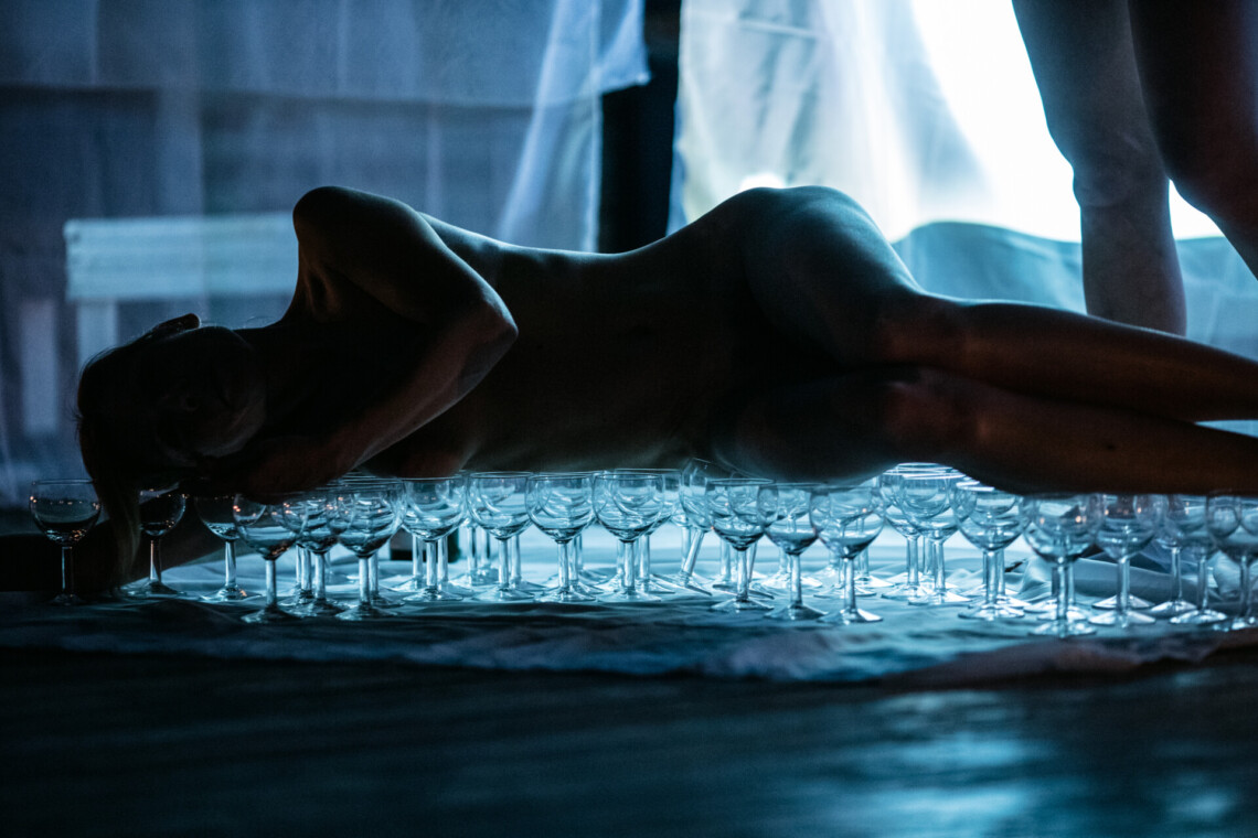 A naked woman lying on many wine glasses, blue background