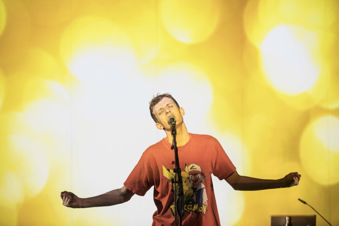 A man in a hoodie stands next to a microphone on a stand, his arms are spread out to the side