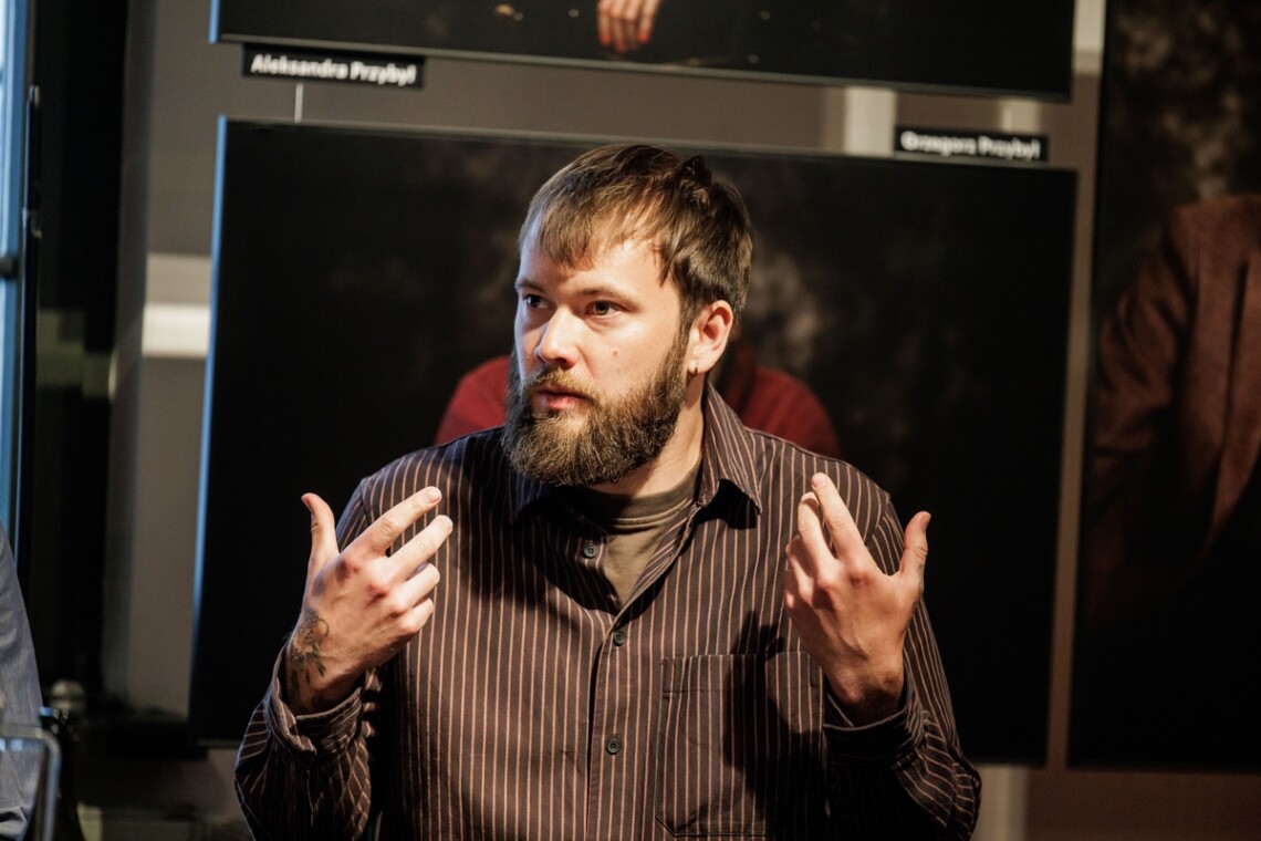 Kacper Lemiesz ubrany w czarną koszule w pionowe paski. Mężczyzna trzyma uniesione dłonie na wysokości barków, wnętrzem skierowane ku sobie.