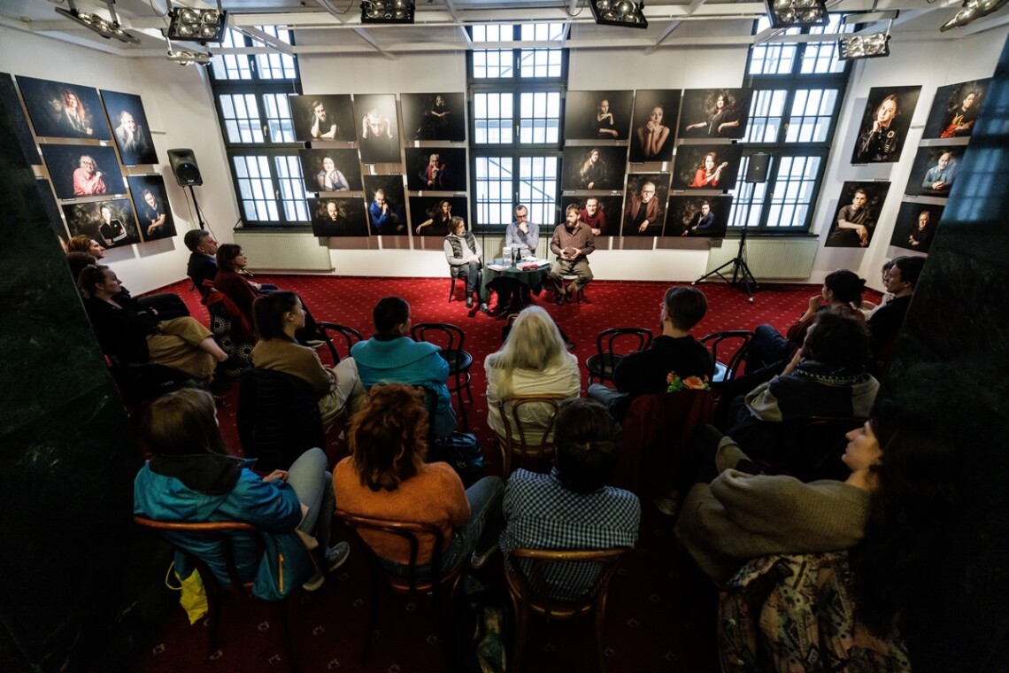 Trzech mężczyzn siedzi na krzesłach, przed nimi stoi okrągły stolik. Za mężczyznami widoczne na ścianie zdjęcia portretowe aktorów Teatru Śląskiego. Przed mężczyznami na krzesłach siedzą widzowie. Ujęcie na wprost zza placów widzów.