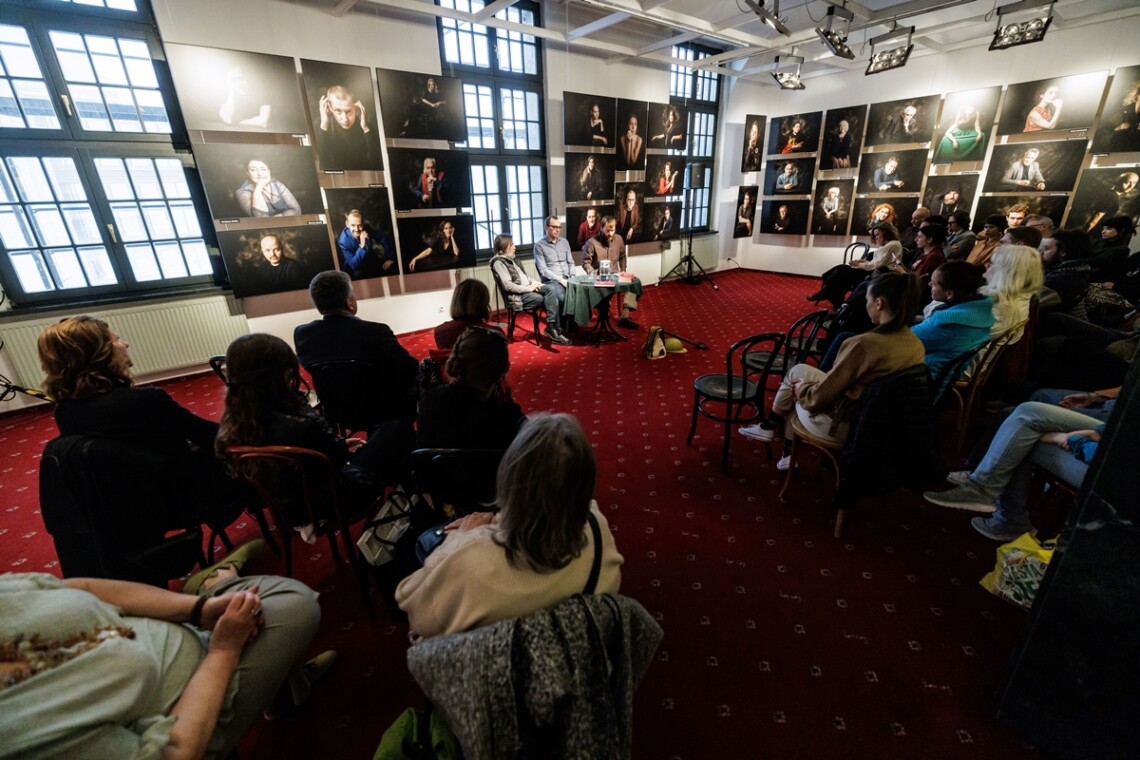 Trzech mężczyzn siedzi na krzesłach, przed nimi stoi okrągły stolik. Za mężczyznami widoczne na ścianie zdjęcia portretowe aktorów Teatru Śląskiego. Przed mężczyznami na krzesłach siedzą widzowie. Ujęcie pod kątem.