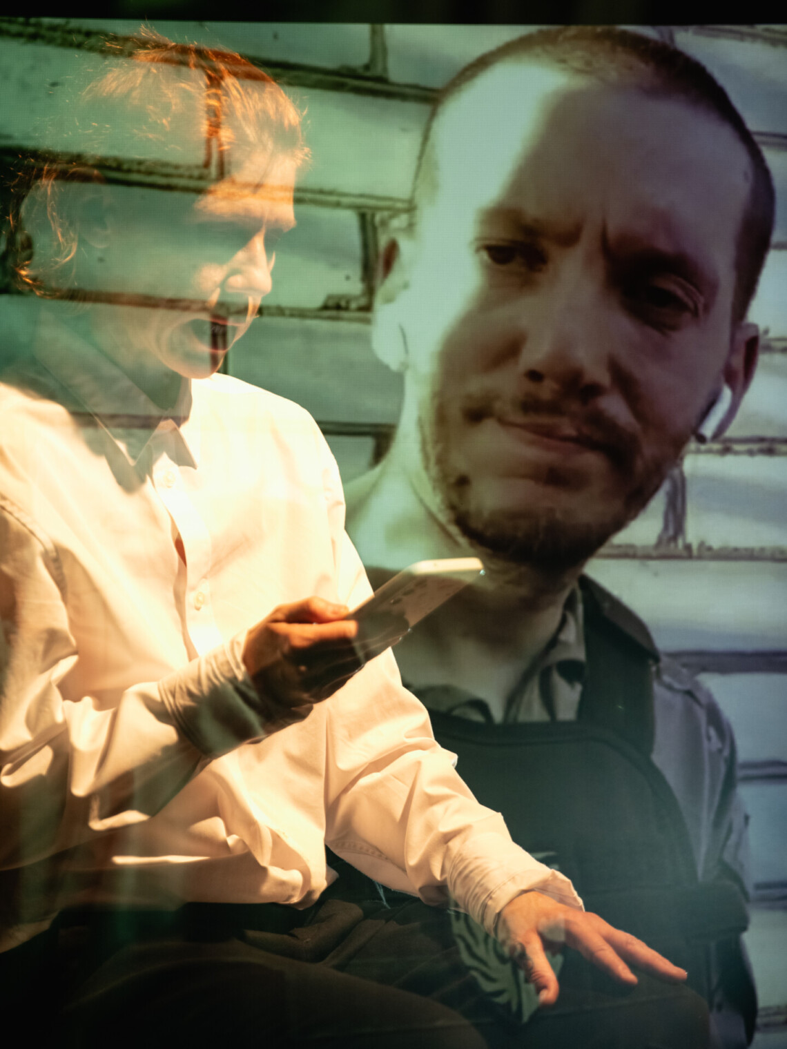 A transparent-looking person sits with a phone in his hand, next to this person a man's figure is displayed on the wall