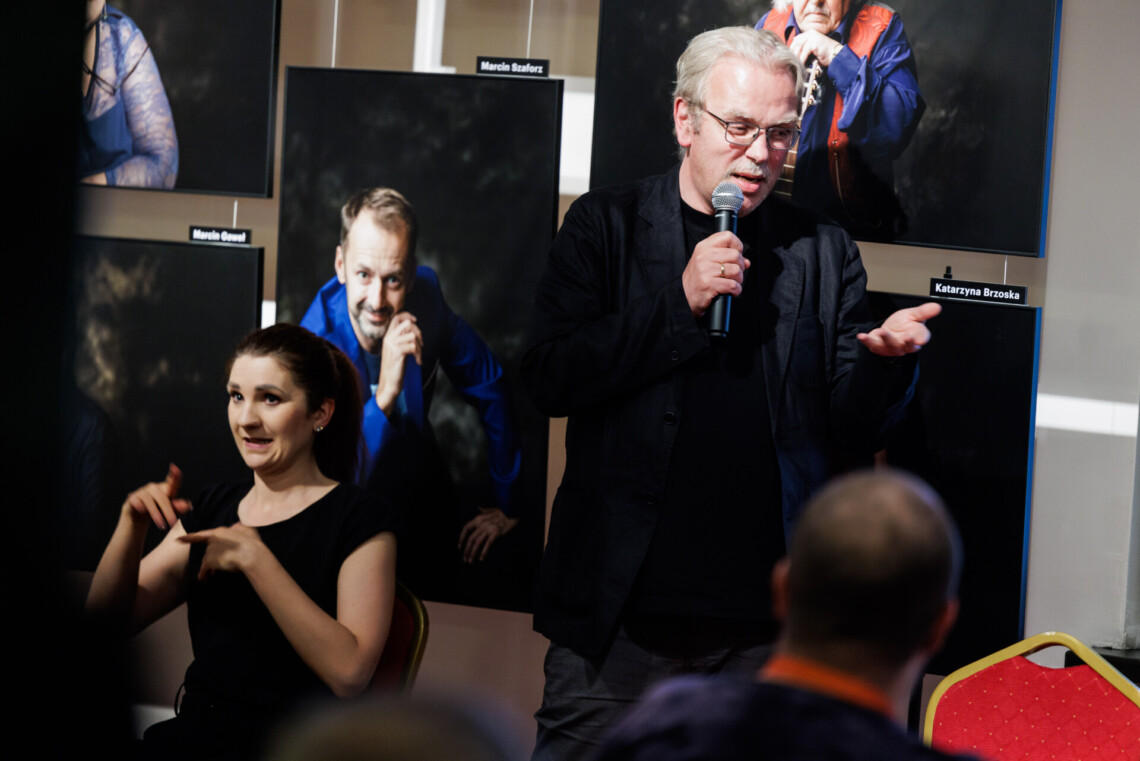 Jacek Wakar stoi z mikrofonem w dłoni Obok niego siedzi tłumaczka Polskiego Języka Migowego w trakcie pracy. W tle widoczne zdjęcia aktorów Teatru Śląskiego.