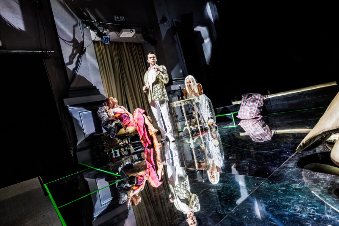 W oddali trzy osoby - kobieta ubrana w różowo -pomarańczową sukienkę w kratę siedzi na krześle, obok niej stoi mężczyzna ubrany w złotą marynarkę i białe spodnie, obok przy złotym okrągłym stoliku siedzi kobieta w srebrnym stroju. Za nimi złota kotara. Na podłodze lustrzane kafle, w której odbijają się występujące postacie.