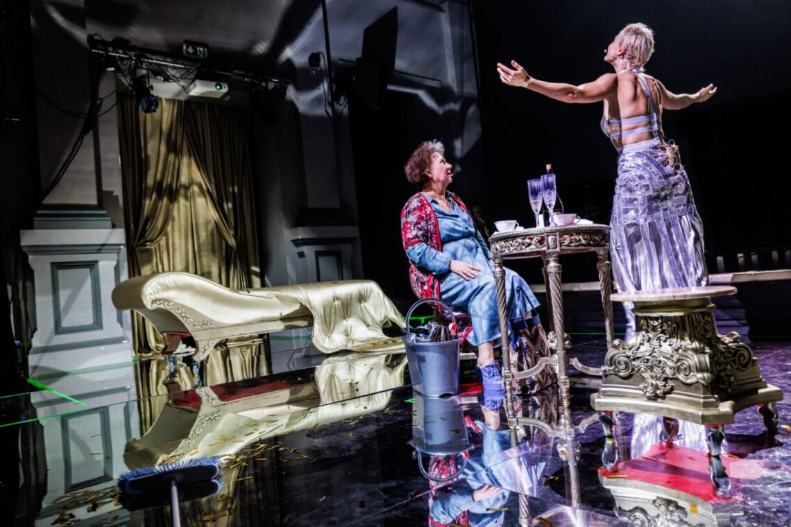 Dwie kobiety, od lewej starsza kobieta w niebieskiej sukience i kwiecistej podomce siedzi na krześle. Naprzeciwko niej stoi młoda kobieta z uniesionymi dłońmi. Ubrana jest w srebrną, długą spódnice oraz srebrny biustonosz. Między kobietami okrągły, złoty stoliku z dwoma kieliszkami do szampana