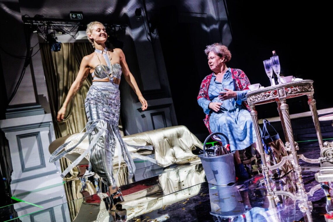 Dwie kobiety, od lewej młoda kobieta ubrana w srebrną, długą spódnice oraz srebrny biustonosz przechadza się po scenie. Po prawej stronie starsza kobieta w niebieskiej sukience i kwiecistej podomce siedzi na krześle przy okrągłym, złotym stoliku, przed nią na podłodze stoi plastikowe wiadro