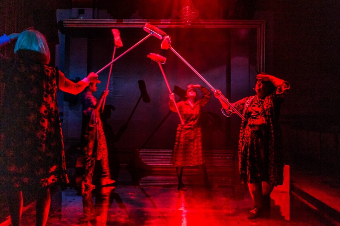 Cztery postacie z wyciągniętymi ku górze drewnianymi miotłami, które stykają się szczotkami w powietrzu. Scena oświetlona czerwonym światłem.