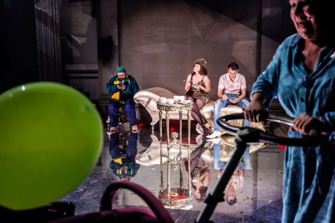 Na pierwszym planie rozmazana postać kobiety w średnim wieku ubranej w niebieską sukienkę, ręce opiera o uchwyt dziecięcego wózka. W tle trzy postacie. Mężczyzna siedzący na fotelu oraz kobieta i mężczyzna na złotym szezlongu. W lustrzanej podłodze odbijają się postacie oraz rekwizyty.