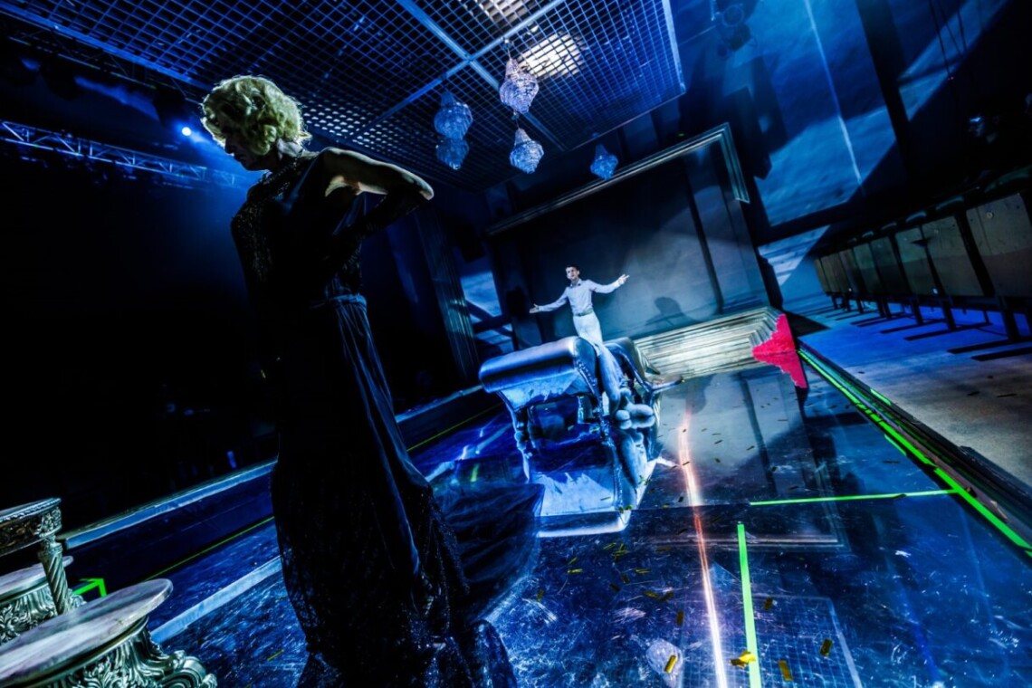 Na pierwszym planie postać stojąca w mroku, ubrana w długą suknię, jej ręce oparte są na boku. Na drugim planie w oddali stoi mężczyzna z rozpostartymi ramionami. W tle elementy dekoracji. Na scenę pada niebieskie światło. Podłoga sceny lustrzana, w które odbijają się aktorzy oraz rekwizyty.