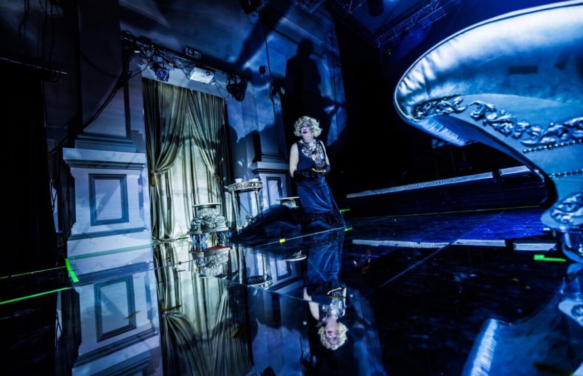 W oddali klęczy mężczyzna ucharakteryzowany na kobietę. Ubrany w czarną, długą sukienkę i blond perukę. Za nim znajdują się złote rekwizyty: stolik i taboret oraz złota kotara. Scena oświetlona niebieskim światłem. Podłoga lustrzana, w której odbija się postać oraz rekwizyty.