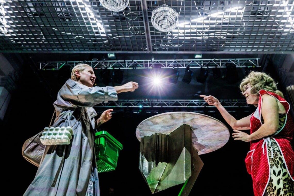 Dwie kobiety stoją na przeciwko siebie z wyciągniętymi prawymi dłońmi, które wskazują w swoim kierunku. Kobieta po lewej ubrana w szary płaszcz, srebrną torebkę, na lewej dłoni przewieszony zielony koszyk sklepowy. Kobieta po prawej ubrana w lamparcią sukienkę i czerwono biały fartuch, na głowie ma założoną blond perukę. Nad nimi sufit ze srebrną kratką i kryształowe żyrandole.