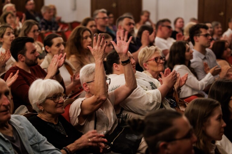 Publiczność siedząca na widowni. Osoby biją brawa, część osób bije brawa w języku migowym, czyli z uniesionymi dłońmi w powietrzu.