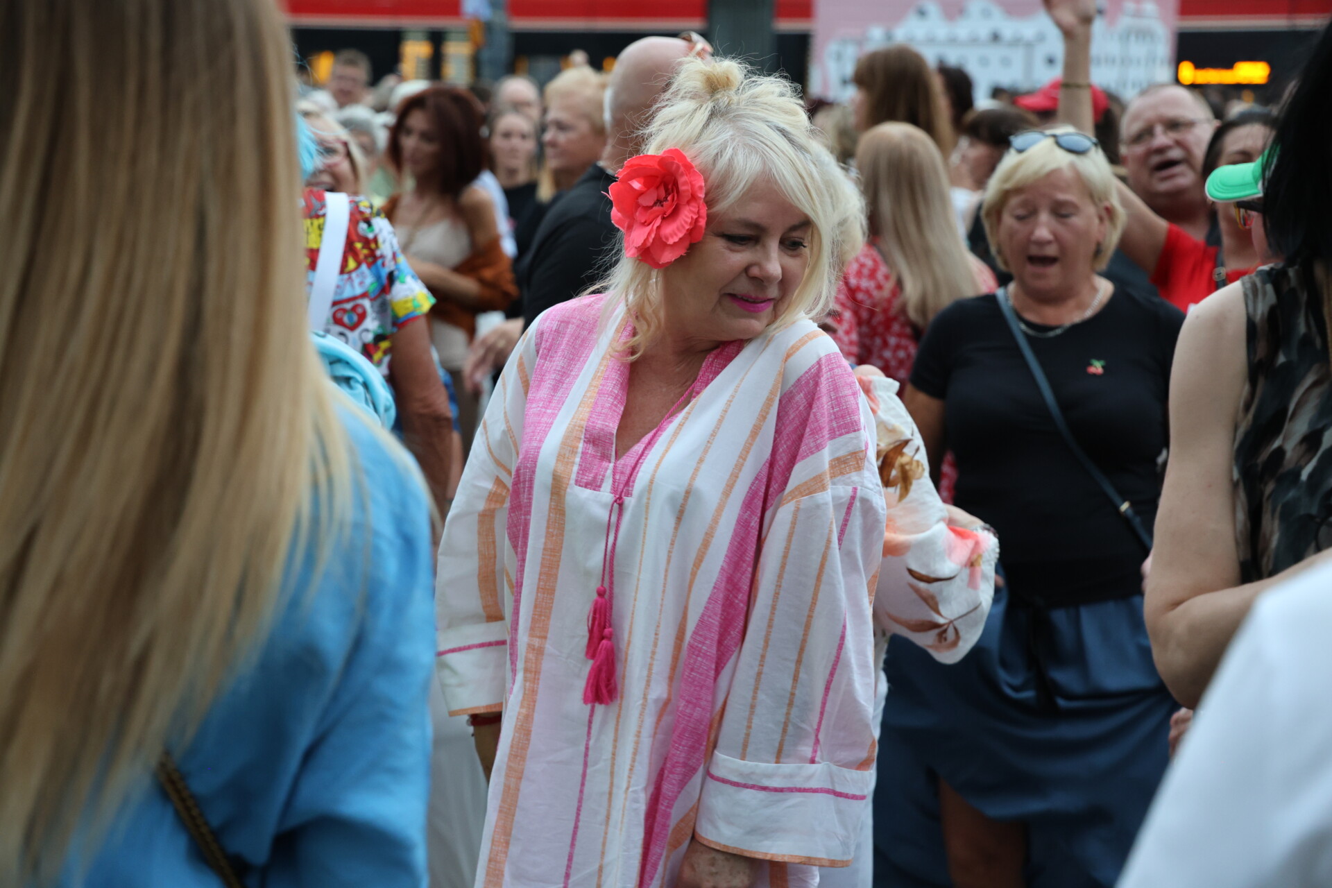 W centrum kobieta z czerwonym kwiatem wpiętym we włosy, wokół niej inne osoby