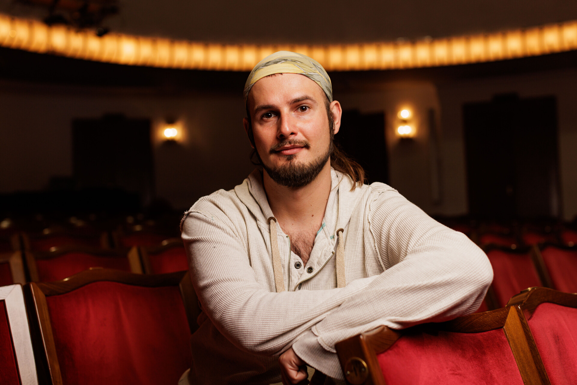 Zdjęcie portretowe Rafała Domagały - mężczyzna w chuście na głowie siedzący w czerwonym fotelu widowni, opiera się o oparcie fotela z rzędu przed, patrzy w obiektyw