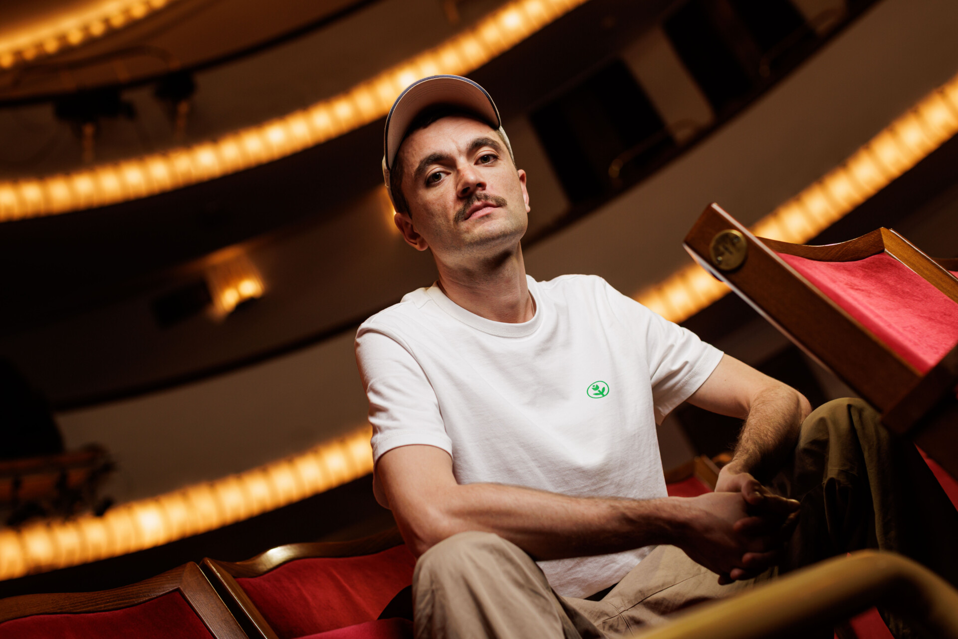 Zdjęcie portretowe Huberta Sulimy - mężczyzna z czapką z daszkiem siedzący na brzegu fotelu widowni, patrzy w obiektyw, ujęcie od dołu