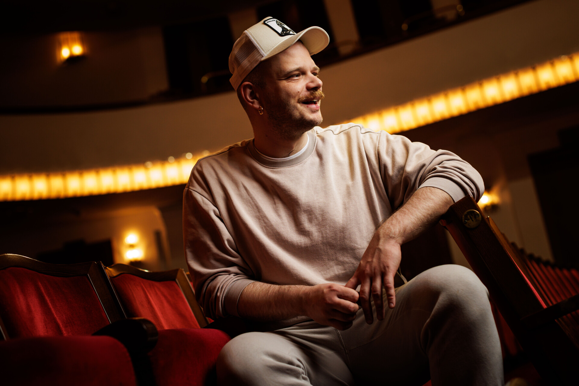 Zdjęcie portretowe Jędrzeja Piaseckiego - mężczyzna z czapką z daszkiem siedzący na brzegu fotelu widowni, patrzy w bok, ujęcie od dołu