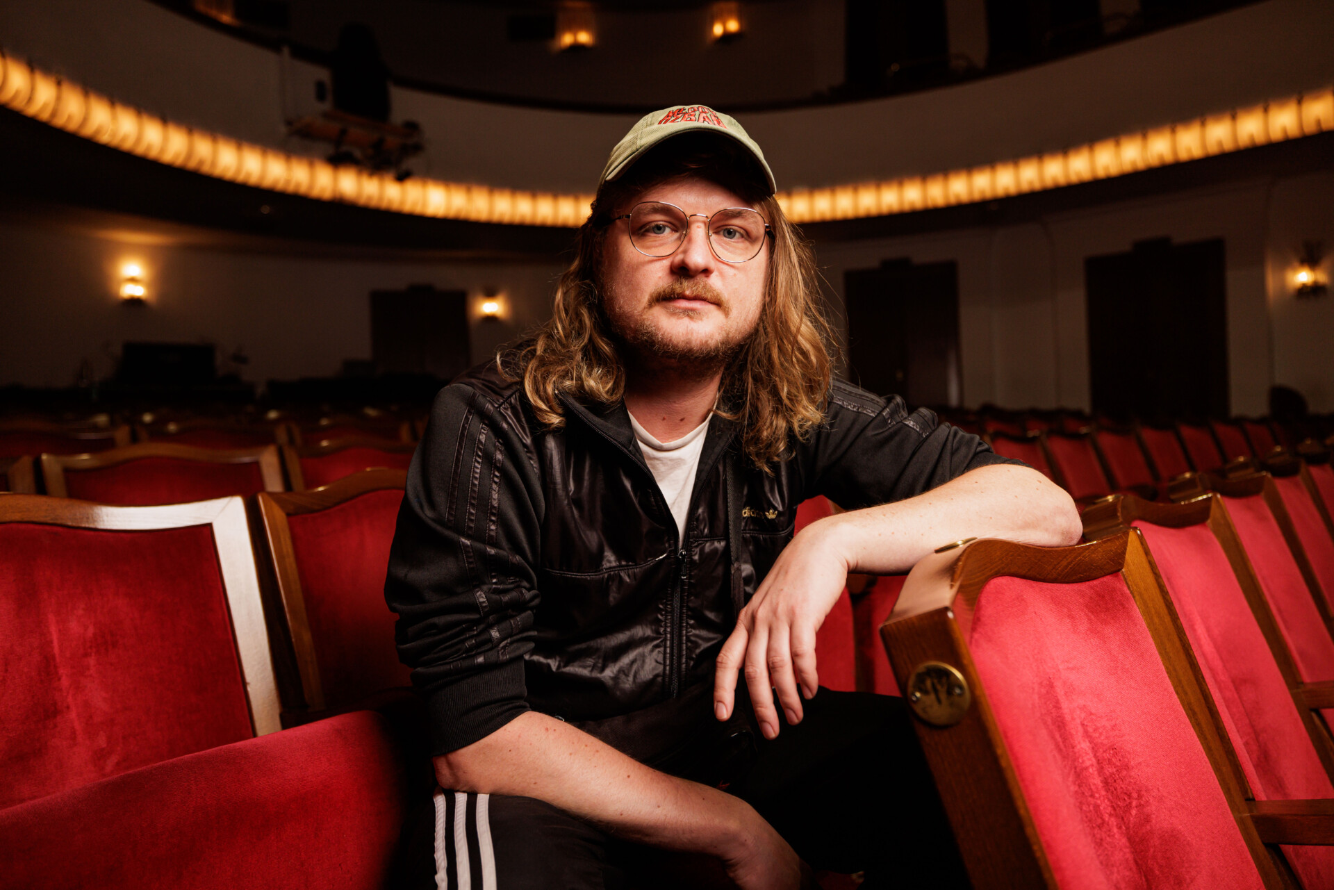 Zdjęcie portretowe Huberta Sulimy - mężczyzna z czapką z daszkiem w okularach siedzący w czerwonym fotelu widowni, opiera się łokciem o oparcie fotela z rzędu przed, patrzy w obiektyw