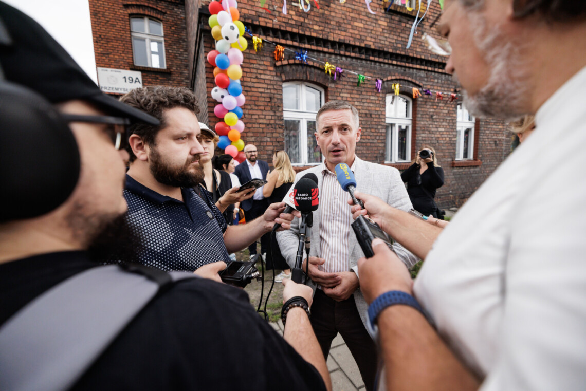 Piotr Kuczera udzielający wywiadu, przed nim stoją dziennikarze z mikrofonami i dyktafonami, za nim budynek z czerwonej cegły - śląski familok