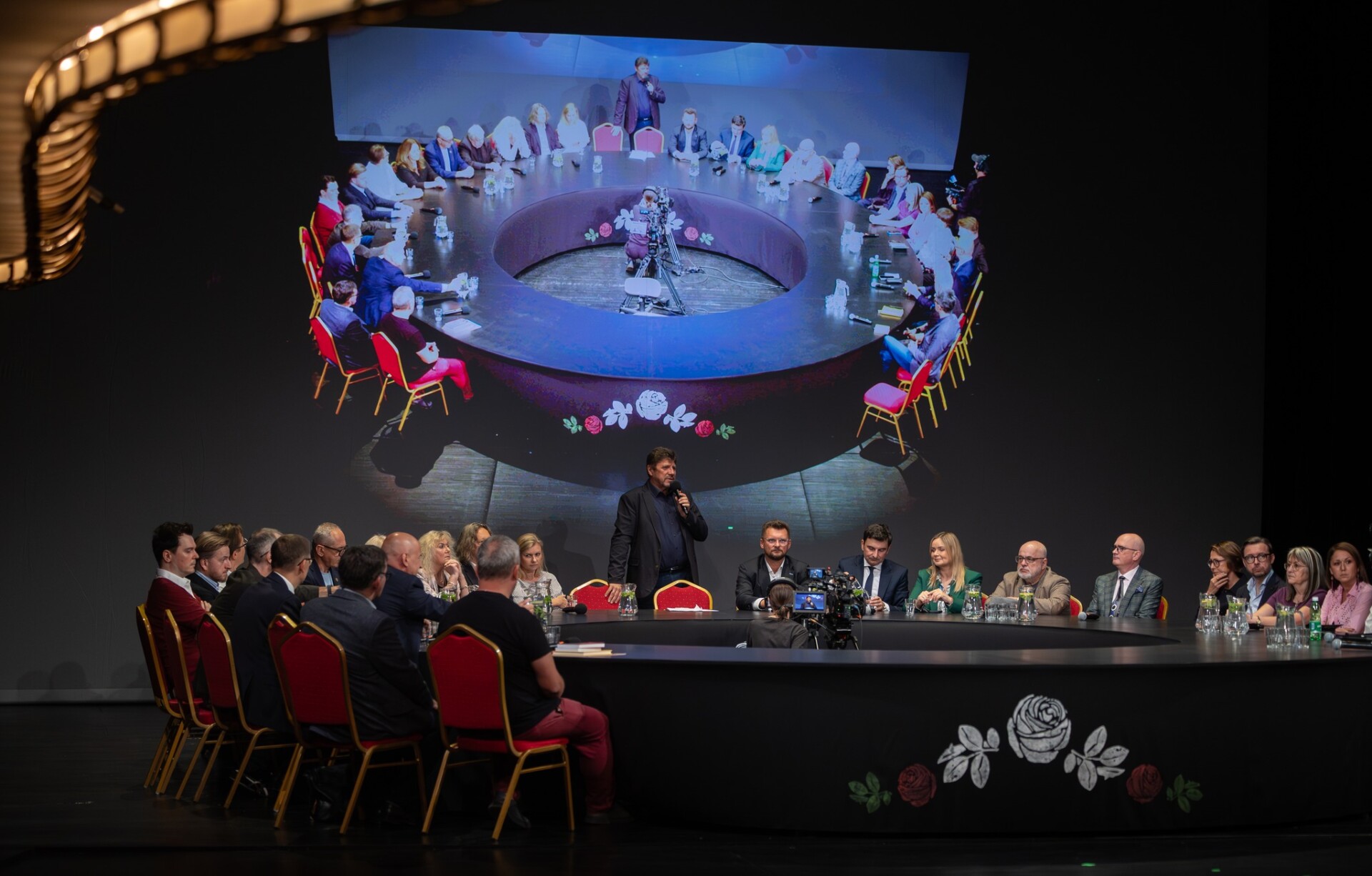 Osoby siedzące wokół czarnego, okrągłego stołu, Na szczycie stołu stoi Dyrektor Robert Talarczyk i przemawia do mikrofonu, za nim na ekranie transmisja spotkania widziana od góry