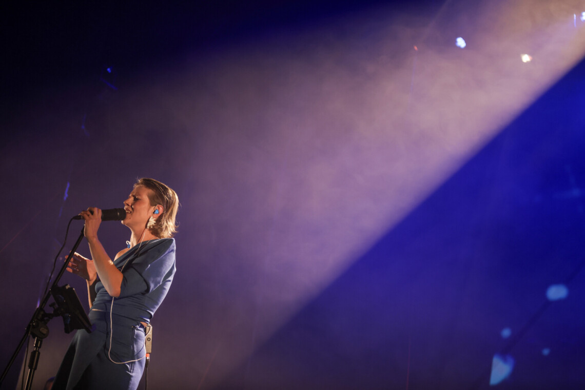 Piosenkarka śpiewajaca do mikrofonu z zamkniętymi oczami, jej postać zajmuje jedną szóstą zdjęcia w lewym dolnym rogu, pozostała część zdjęcia to padające jasne i niebieskie światło