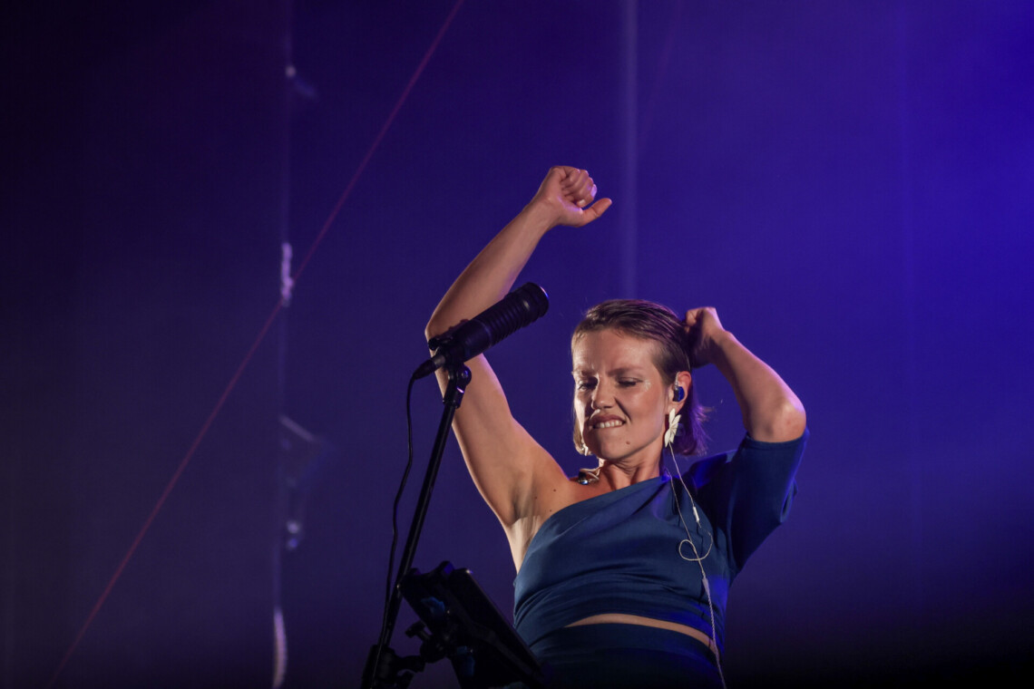 Piosenkarka z uniesionymi do góry rękami, wygląda jakby tańczyła, ma zamknięte oczy i zaciśnięte zęby, tło oświetlone na niebiesko