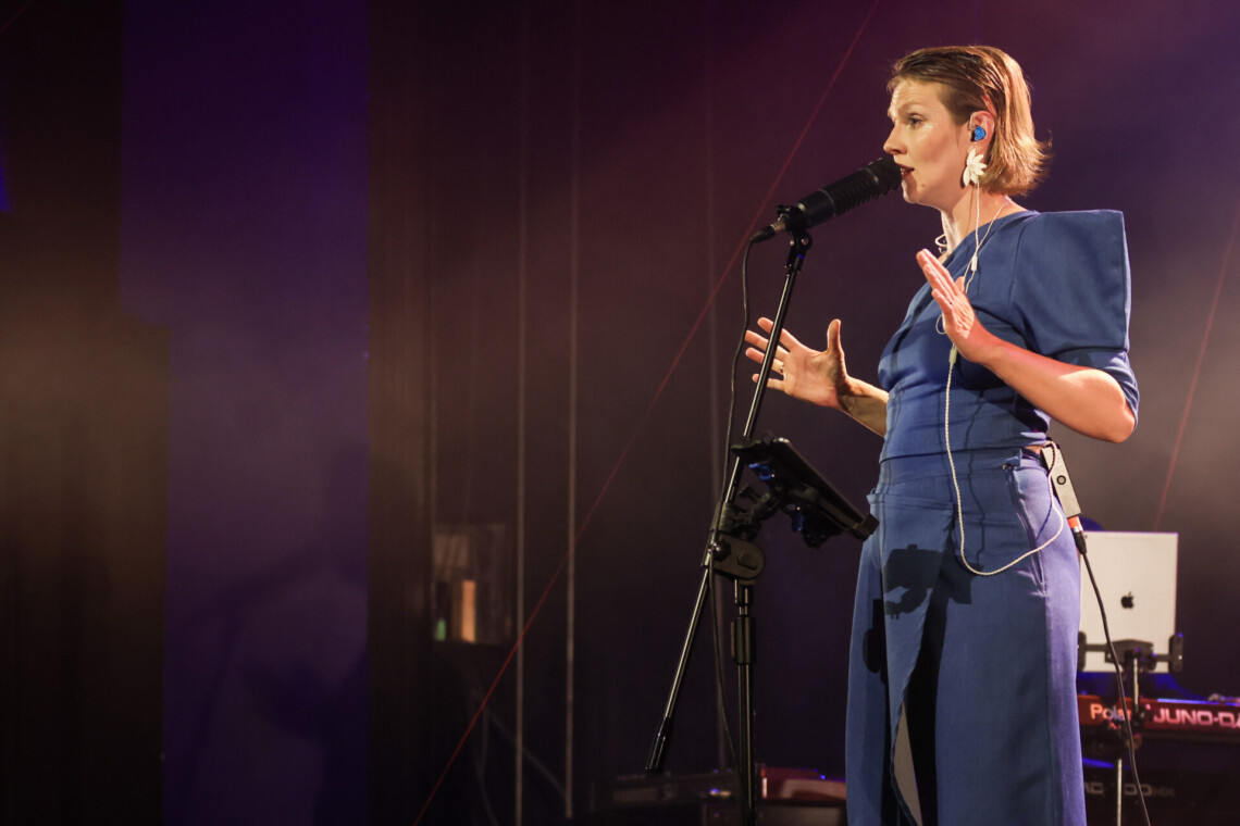 Piosenkarka stojąca przy mikrofonie na statywie, zgięte ręce z rozłożonymi palcami dłoni trzyma na wysokości kratki piersiowej, ujęcie z boku