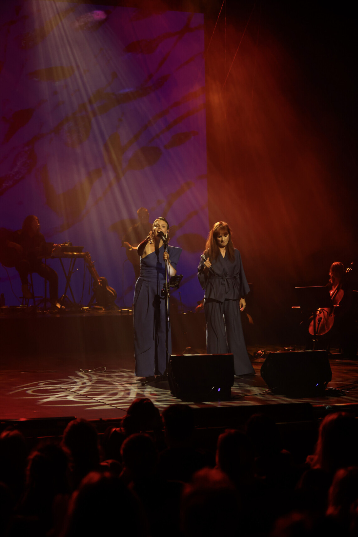 Anna Bratek oraz Agnieszka Radzikowska w niebieskich sukienkach na scenie, scena jest oświetlona na czerwono, na podłodze sceny widoczne są mazie jasnego światła, ujęcie z oddali