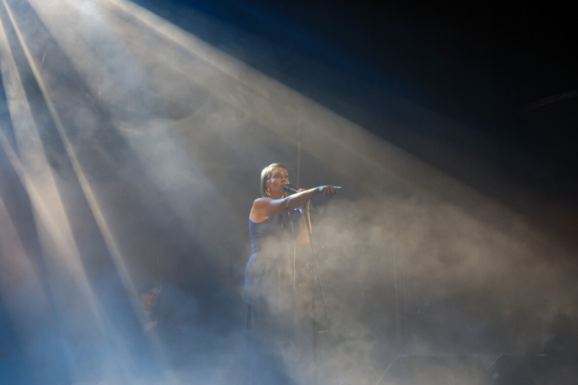 Piosenkarka przy mikrofonie na statywie, prawą rękę ze wskazującym palcem ma wysuniętą do przodu, scena spowita jest mgłą, z góry pod kątem pada światło, kobieta znajduje się w jego środku