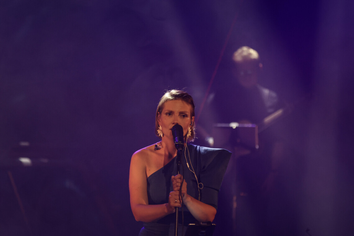 Piosenkarka trzyma dłońmi statyw mikrofonu, w tle widoczny muzyk; ujęcie na wprost