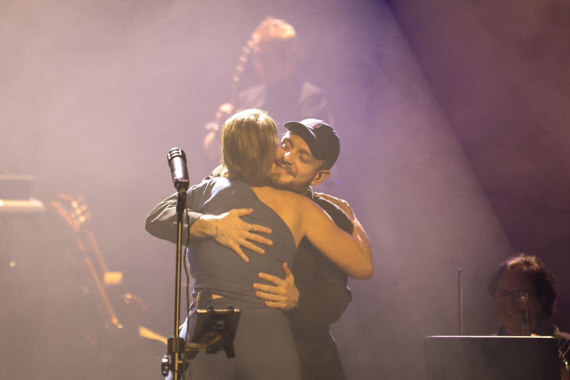 Piosenkarka przytulająca się z muzykiem, widoczne są plecy kobiety, a twarz mężczyzn w czapce z daszkiem zza jej ramienia