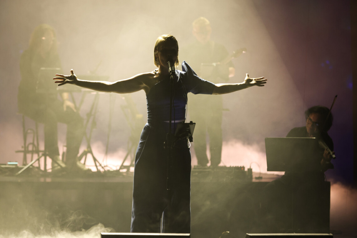 Piosenkarka stojąca z rozpostartymi na bok rękami, za nią muzyka, scena oświetlona jasnym światłem, kobieta stoi w cieniu