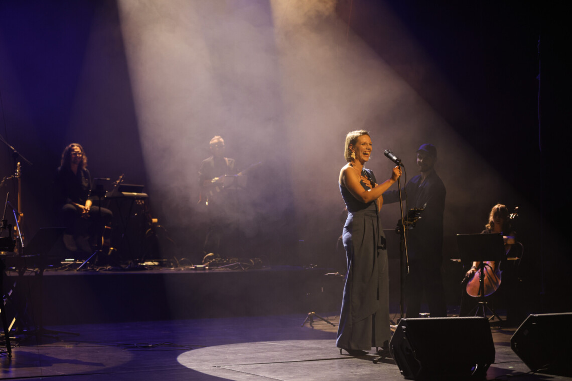 Piosenkarka i muzycy na scenie, w centrum kobieta przy statywie śmieje się opierając dłoń na klatce piersiowej, z góry pada na nią jasne światło tworząc na podłogę okrąg; ujęcie z boku