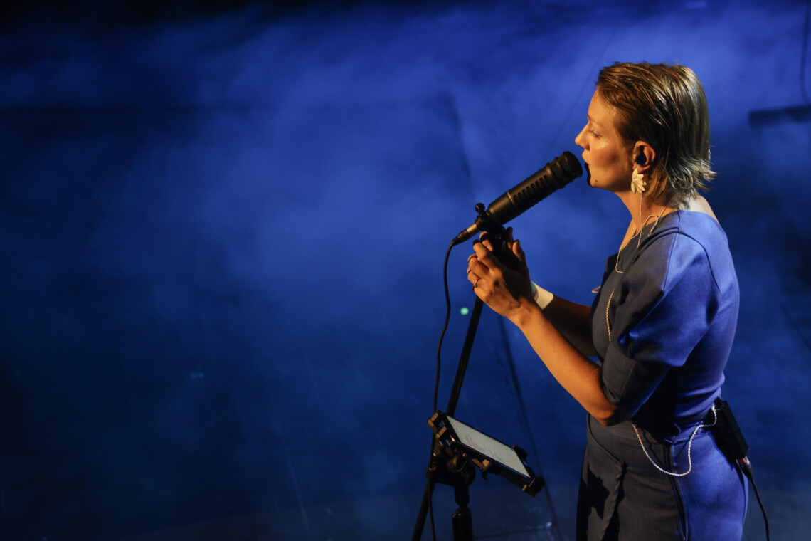 Piosenkarka przy mikrofonem na statywie, który obejmuje dłońmi, ujęcie z boku, od góry, scena oświetlona na niebiesko