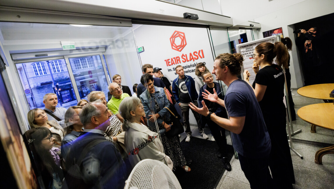 Aktor Bartosz Błaszczyński przemawia do grupy zwiedzających. Jego dłonie są uniesione na wysokości klatki piersiowej, sylwetka zwrócona bokiem do obiektywu. Po jego prawej stronie stoi tłumaczka Polskiego Języka Migowego w trakcie pracy.