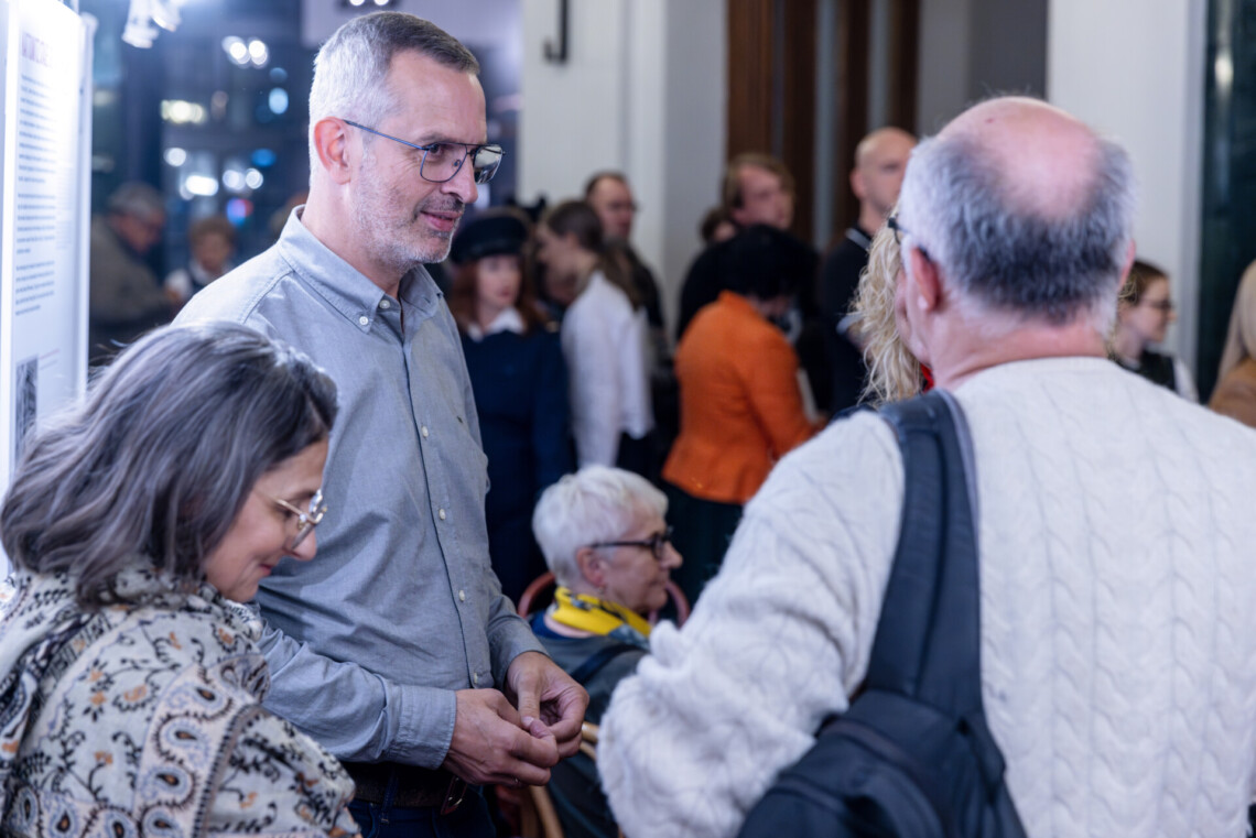 Na pierwszym planie trzy osoby stojące w foyer, za nimi rozmazane postacie osób stojących w kolejce