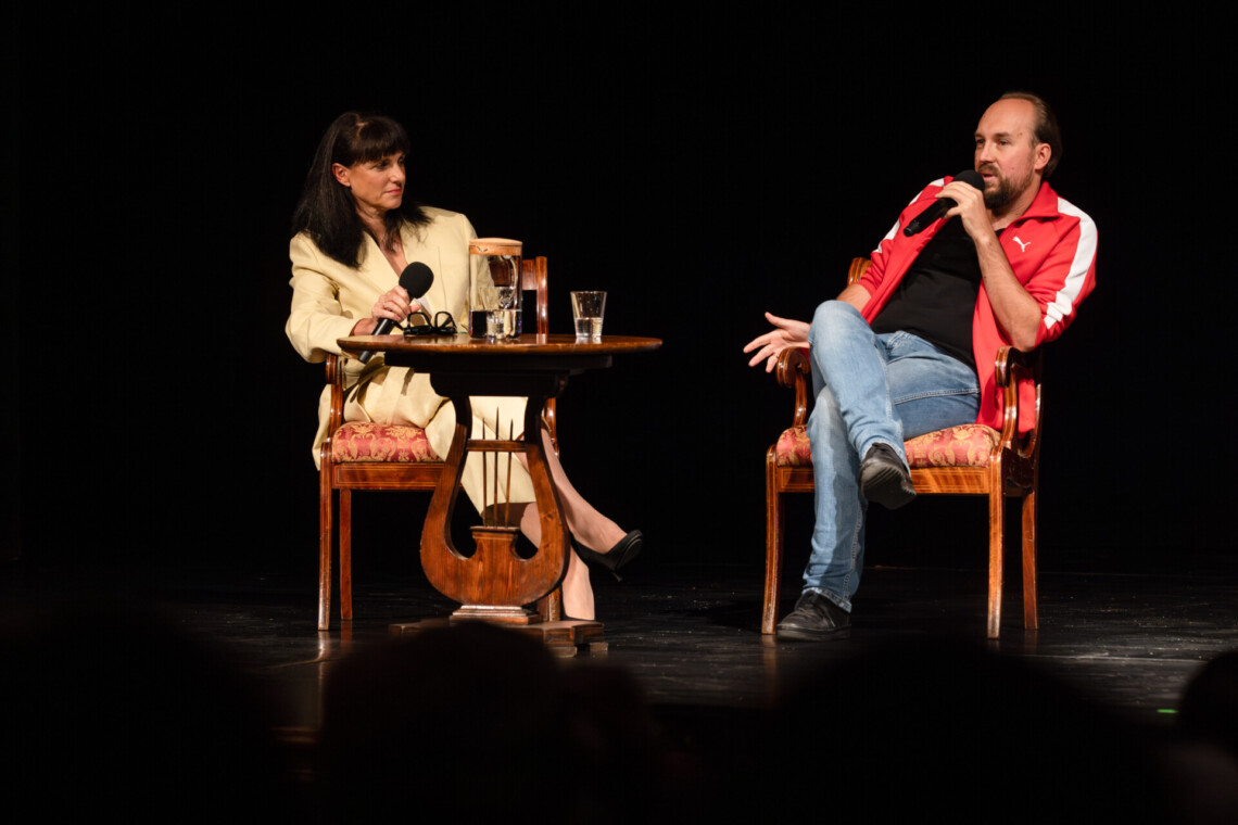 Przy okrągłym stoliku siedzą Anna Dudzińska oraz Zbigniew Rokita przemawiający do mikrofonu; tło za nimi czarne, na pierwszym planie kontury czubki głów osób siedzących na widowni