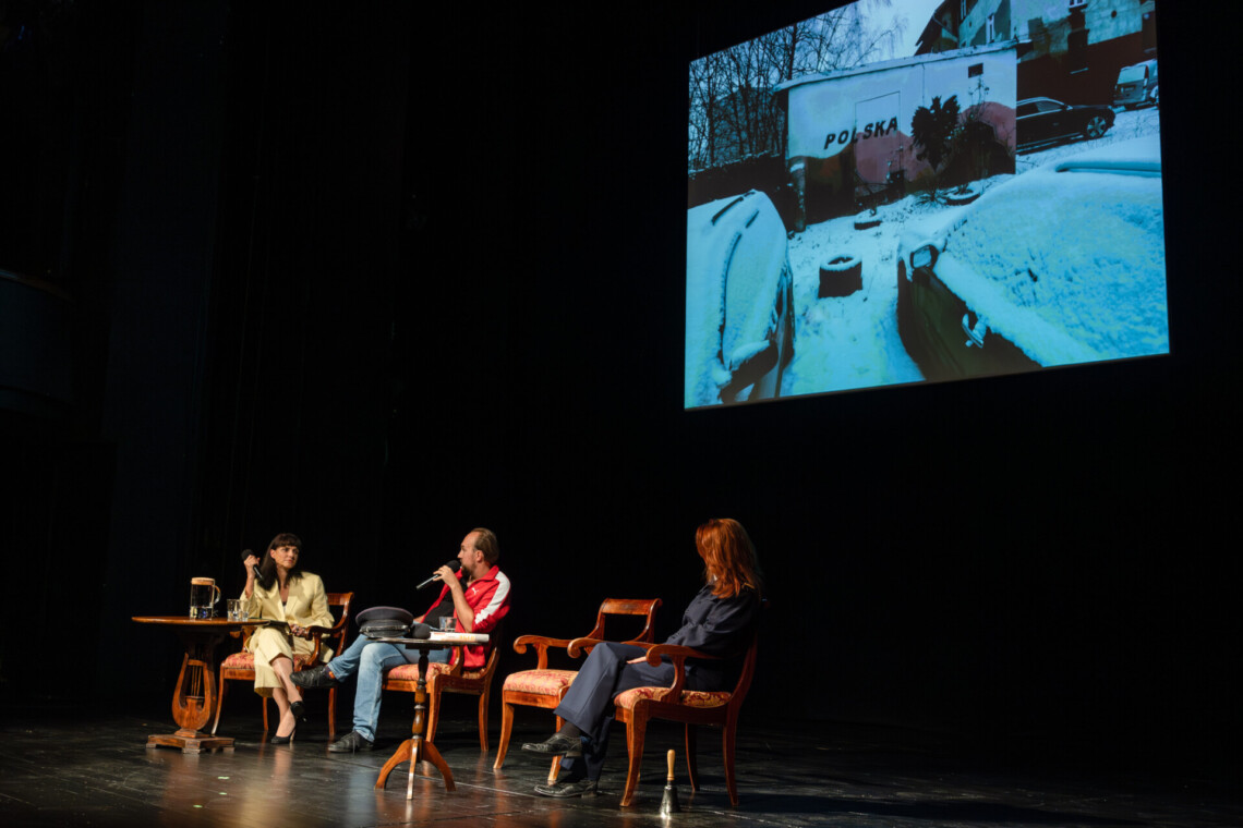 Na scenie przy dwóch oddzielnych, okrągłych stołach siedzą trzy osoby, jedno krzesło jest puste, do mikrofonu przemawia Anna Dudzińska, za nimi na ścianie zawieszony ekran z zimowym kadrem, ujęcie pod kątem